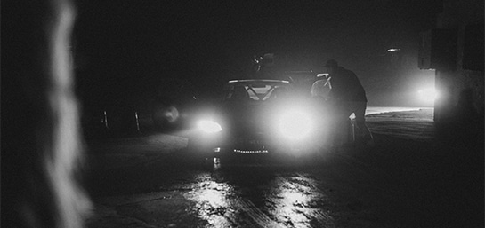 Auto en una vía oscura con las luces encendidas. Imagen a blanco y negro.